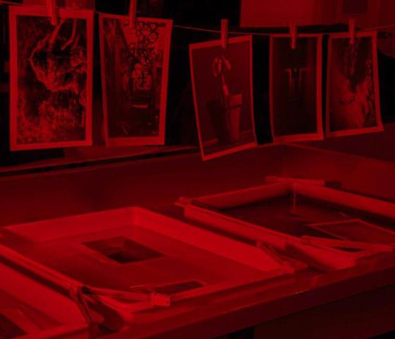 A darkroom lit up in red light with phots hanging on a line and in developing trays